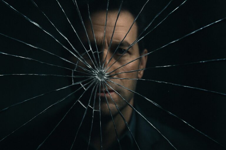 Close-up of a cracked mirror with a man’s face partially reflected through the shards, symbolizing fractured self-reflection.