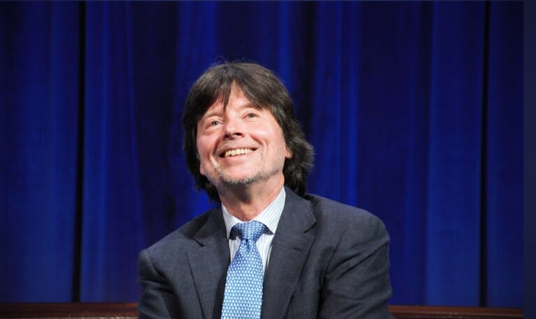Ken Burns smiling on stage in a suit and blue tie against a dark blue curtain background
